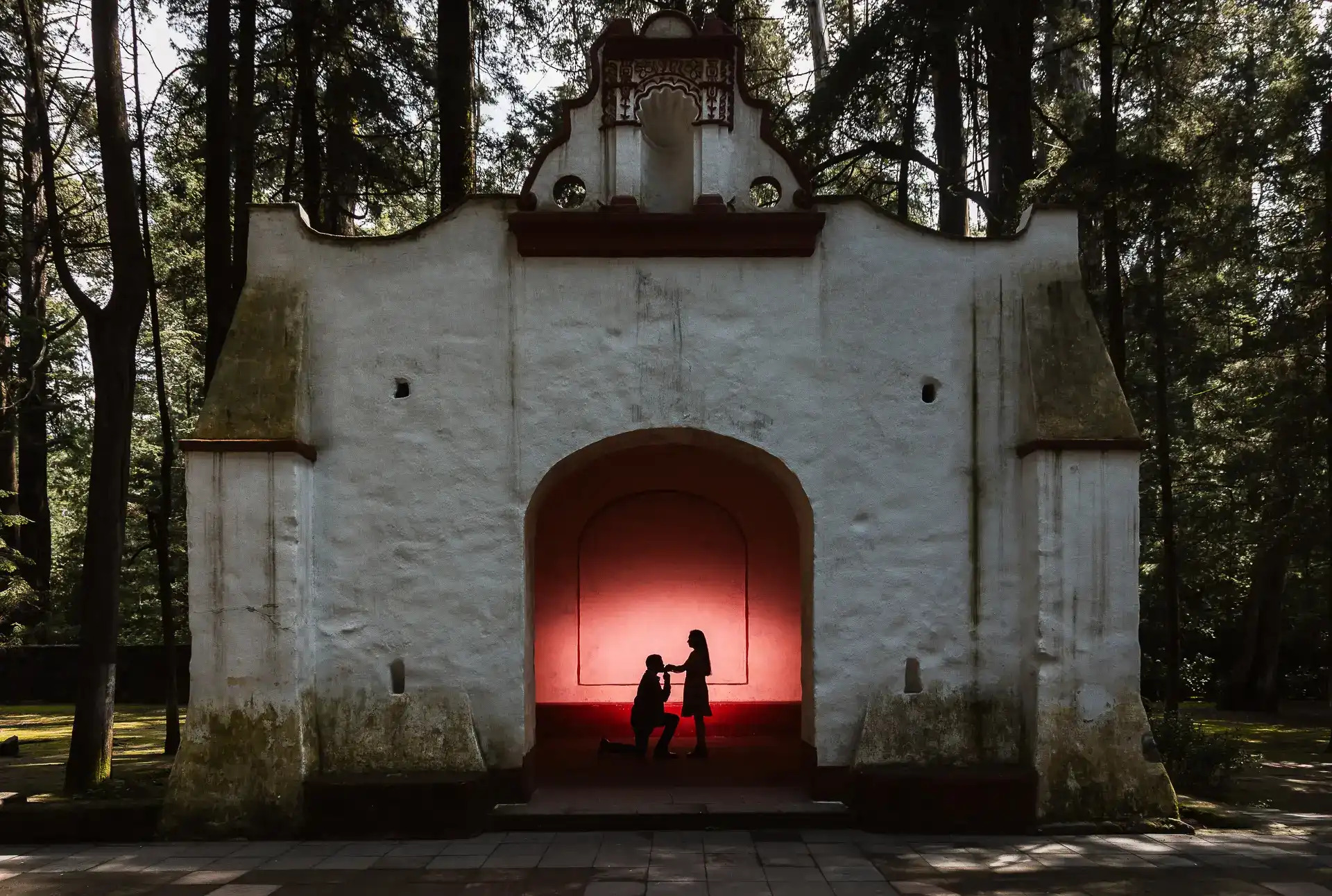 Pedida de mano en silueta bajo arco colonial mexicano con luz roja dramática, fotografía editorial por Salicatus