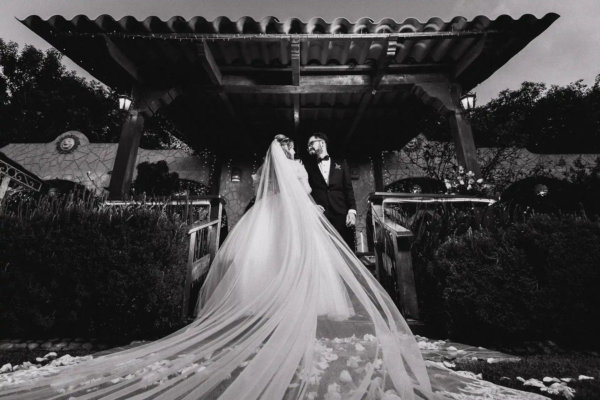 Novios bajo pérgola colonial de noche con velo largo extendido en blanco y negro, fotografía nocturna de boda