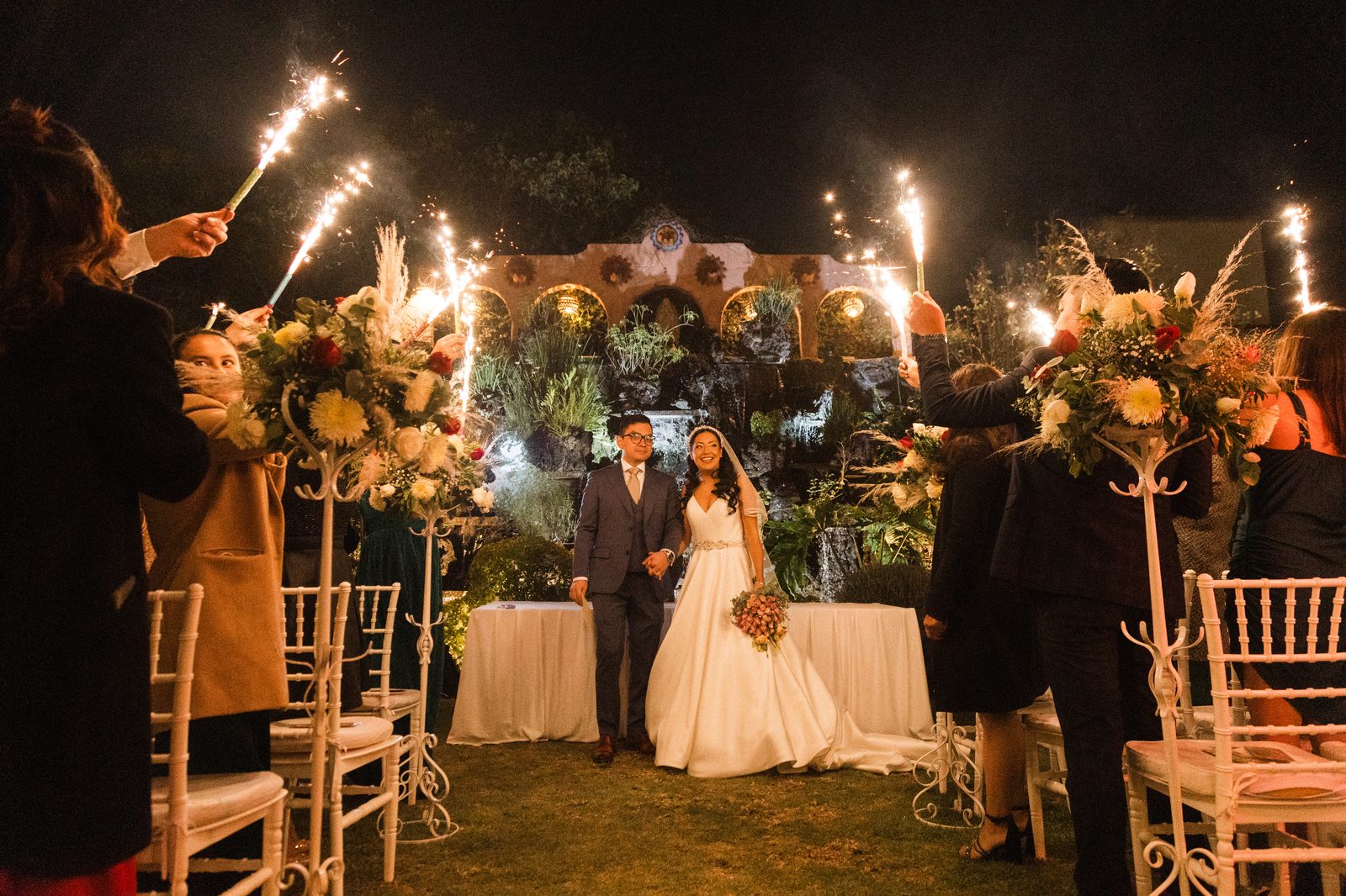 Novios con bengalas y pampas en jardín nocturno con arcos coloniales — boda fotografiada por Salicatus