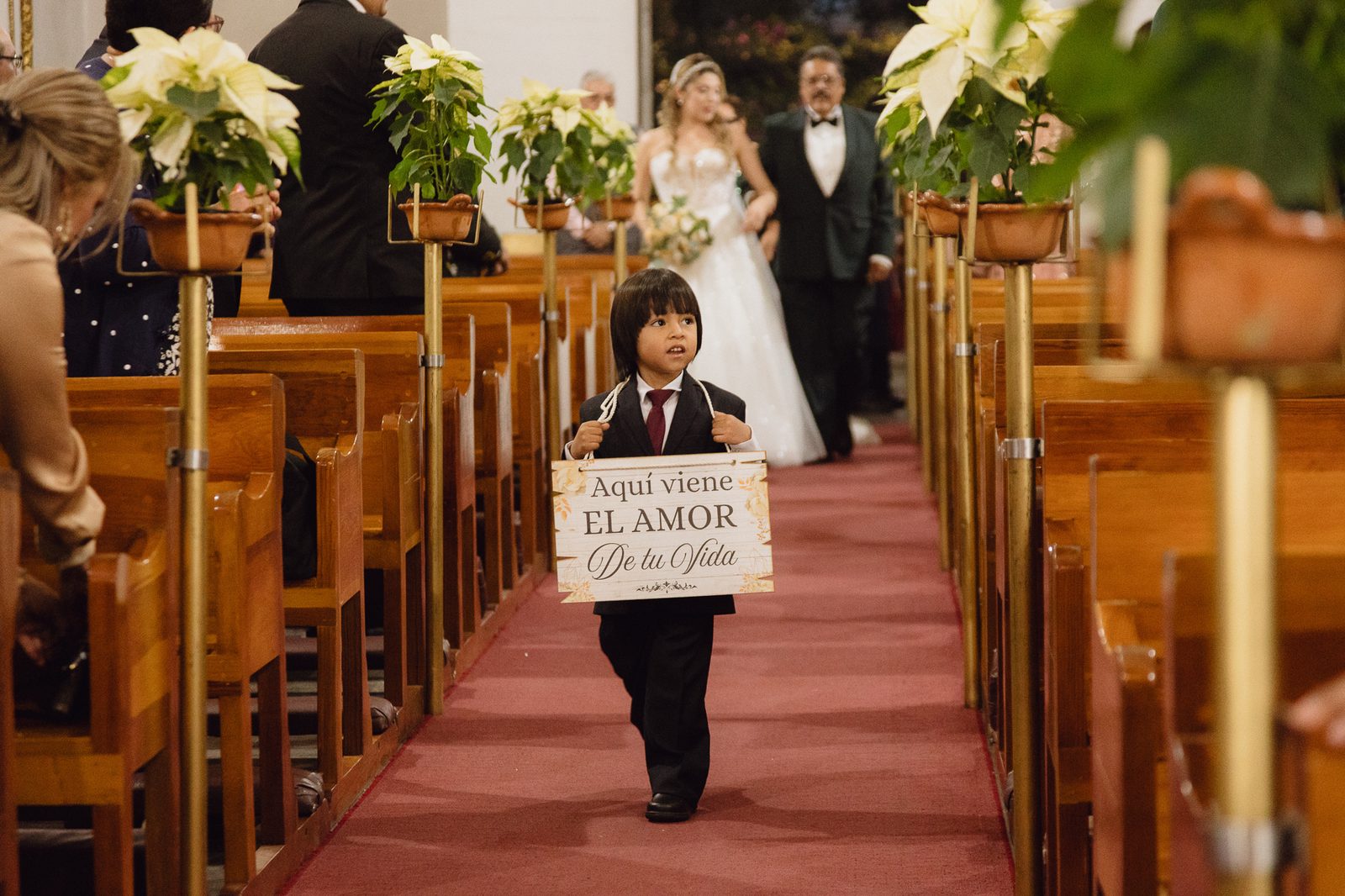 Niño paje cargando cartel 'Aquí viene el amor de tu vida' caminando por el pasillo de la iglesia antes de la novia, fotografía documental de boda — Salicatus