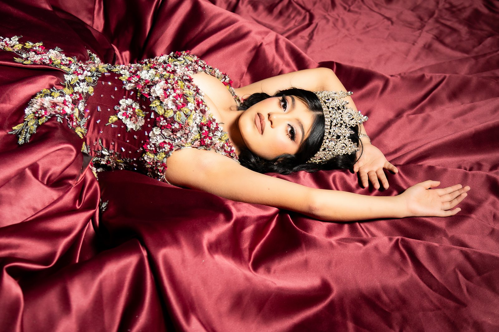 Quinceañera recostada sobre tela roja con vestido floral 3D y corona, sesión Glam editorial estilo Vogue para XV años — Salicatus