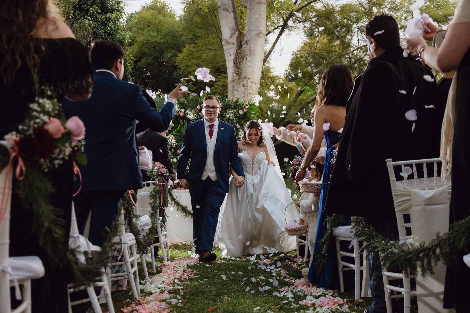 Novios saliendo de ceremonia al aire libre mientras los invitados lanzan pétalos de rosa, momento de celebración en boda al jardín — Salicatus